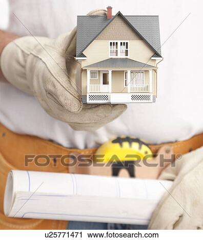Stock Image - Close up of manual worker holding model house and blue print. Fotosearch