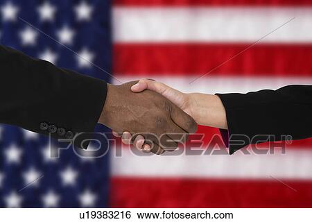 Stock Photograph - Close-up of people shaking hands against American flag. Fotosearch