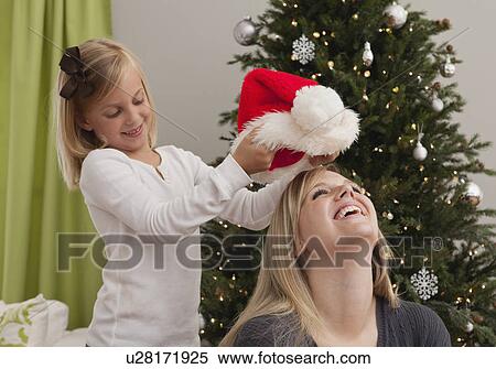 Stock Photography - Daughter (6-7) putting Santa Hat on mother's head in front of Christmas tree. Fotosearch