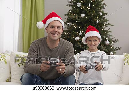 Stock Image - Father and son (8-9) wearing santa hats playing computer games with christmas tree in background. Fotosearch