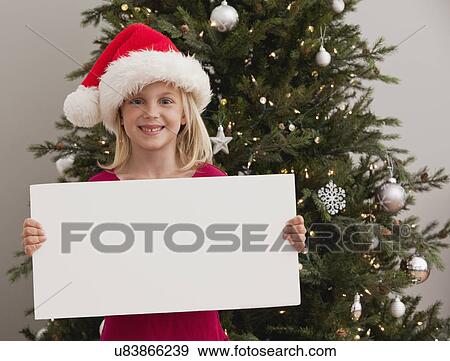 Stock Photo - Girl (-6-7) wearing Santa hat holding blank card in front of christmas tree. Fotosearch