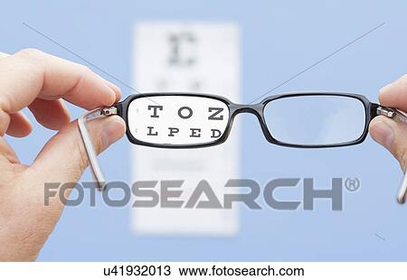 Stock Image - Hands holding glasses in front of eye chart, studio shot. Fotosearch