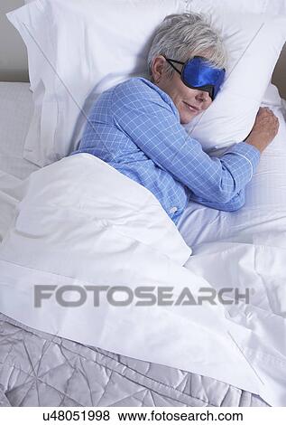 Stock Photo - High angle view of mature woman wearing sleep mask lying in bed. Fotosearch