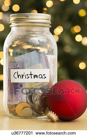Stock Image - Jar filled with money labeled Christmas and Christmas ornaments lying beside. Fotosearch