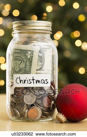 Stock Image - Jar filled with money labeled Christmas and Christmas ornaments lying beside. Fotosearch