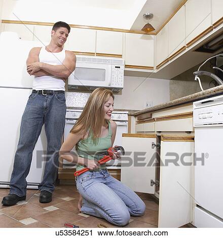 Stock Image - Man watching female plumber fixing kitchen sink. Fotosearch
