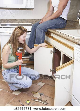 Stock Image - Man watching female plumber fixing kitchen sink. Fotosearch