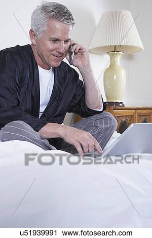Stock Image - Mature man sitting on bed, talking on mobile phone and using laptop. Fotosearch