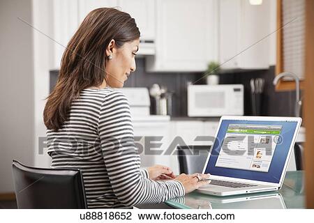 Stock Image - Mature woman using laptop in kitchen. Fotosearch
