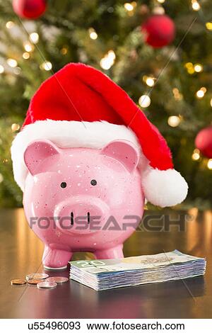 Stock Image - Piggy bank wearing Santa Claus hat placed on table with currency. Fotosearch