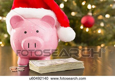 Piggy bank wearing Santa Claus hat placed on table with currency View Large Photo Image Stock Image - Piggy bank wearing Santa Claus hat placed on table with currency. Fotosearch