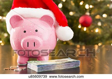 Stock Photo - Piggy bank wearing Santa Claus hat placed on table with currency. Fotosearch