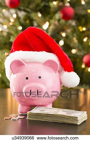 Stock Photograph - Piggy bank wearing Santa Claus hat placed on table with currency. Fotosearch