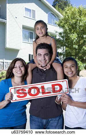 Stock Image - Portrait of family with two girls (8-9, 14-15) holding "Sold" sigh in front of house. Fotosearch