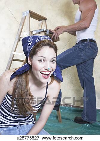 Stock Photo - Portrait of smiling young woman, man painting wall in background. Fotosearch