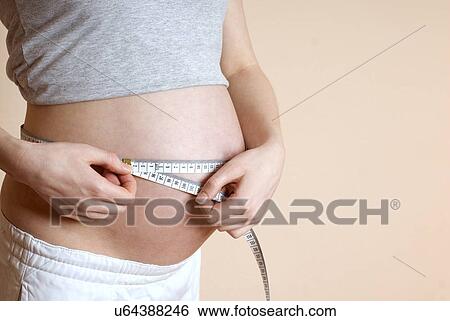 Stock Photograph - Pregnant woman measuring belly with measure tape, mid section. Fotosearch