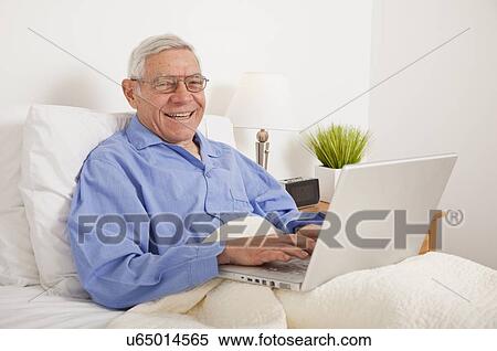 Stock Photography - Senior man using laptop, lying in bed. Fotosearch