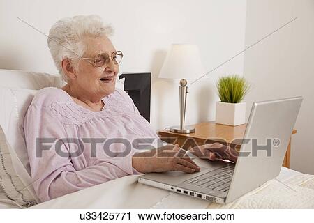 Stock Image - Senior woman in bed, using laptop. Fotosearch
