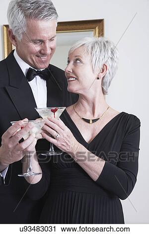 Stock Image - Smiling mature couple in evening wear holding cocktails. Fotosearch