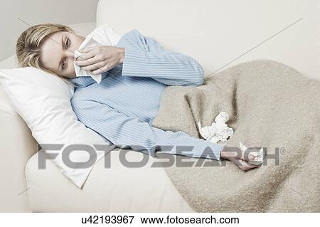 Stock Photo - South Africa, Cape Town, Young woman lying on sofa and blowing nose. Fotosearch