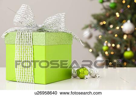 Stock Image - Studio shot of green gift on table with christmas tree in background. Fotosearch