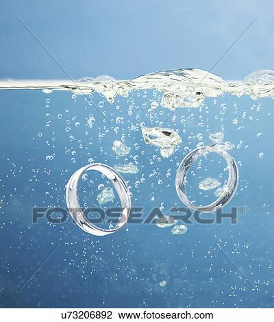 Stock Image - Studio shot of silver wedding rings falling into water. Fotosearch