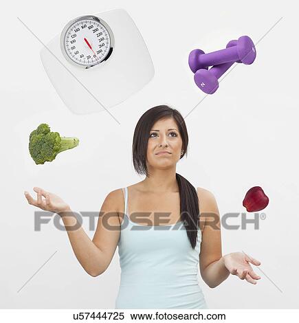 Stock Photography - Studio shot of young woman juggling with weight scale, weights, fruit and vegetable. Fotosearch
