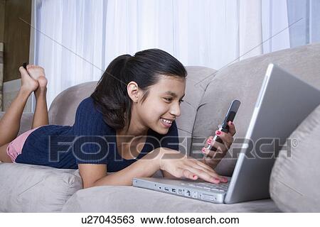 Stock Image - Teenage girl (14-15) using laptop and mobile phone on sofa. Fotosearch