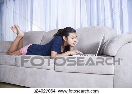 Picture - Teenage girl (14-15) using laptop on sofa. Fotosearch
