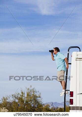 Stock Image - USA, Arizona, mid adult man standing on ladder of mobile home and looking through binoculars. Fotosearch