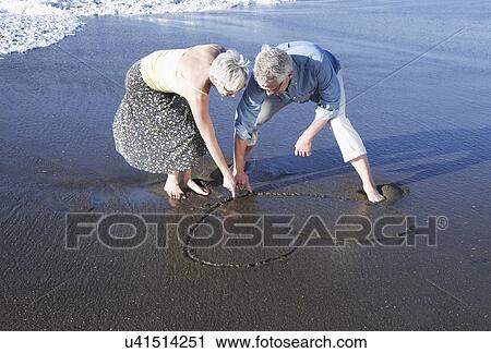 Stock Image - USA, California, Fairfax, Happy mature couple drawing heart on beach. Fotosearch