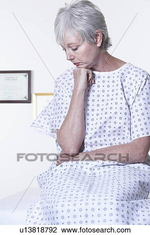 Stock Image - USA, California, Fairfax, Pensive woman in hospital gown sitting in doctor's office. Fotosearch