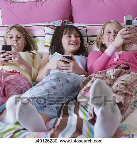 Stock Image - USA California, Fairfax, teenage girls (14-15, 16-17) lying on bed and looking at mobile phones. Fotosearch