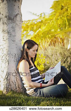 Stock Image - USA, California, Lawndale, Woman sitting under tree reading newspaper. Fotosearch