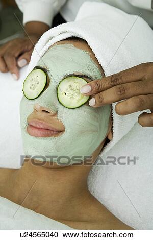 Stock Image - USA, California, Oakland, beautician placing cucumber slice on young woman's eye in spa. Fotosearch