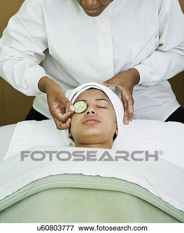 Stock Photo - USA, California, Oakland, beautician placing cucumber slice on young woman's eye in spa. Fotosearch