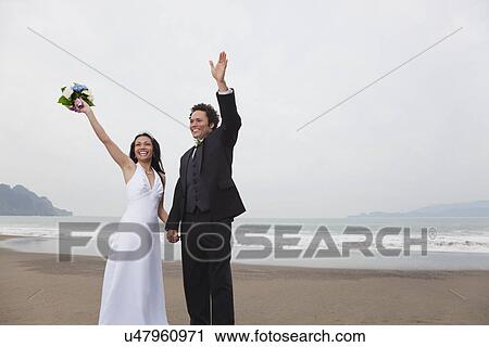 Stock Image - USA, California, San Francisco, Baker Beach, bride and groom on beach waving. Fotosearch
