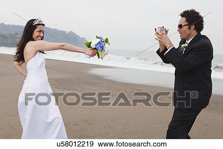 Stock Photo - USA, California, San Francisco, Baker Beach, groom filming bride on beach. Fotosearch