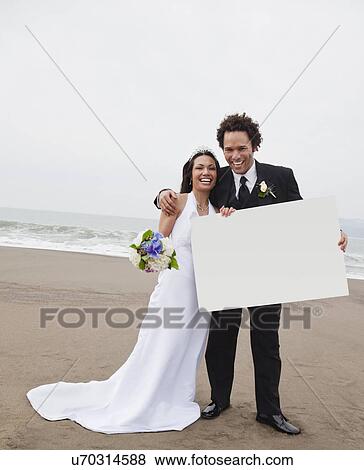 Stock Photo - USA, California, San Francisco, Baker Beach, bride and groom on beach holding placard. Fotosearch