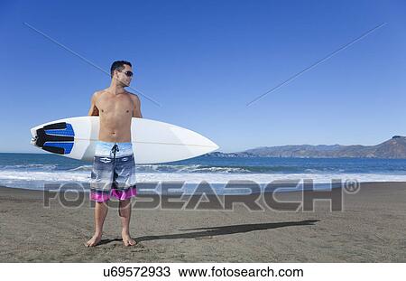 Stock Image - USA, California, San Francisco, man holding surfboard on Baker Beach. Fotosearch