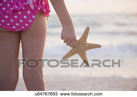 Stock Image - USA, Florida, St. Pete Beach, Rear view of girl (8-9) holding starfish and facing ocean, mid section. Fotosearch