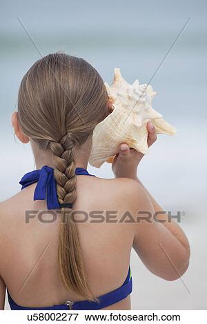 Stock Photo - USA, Florida, St. Pete Beach, Rear view of girl (8-9) listening to conch shell on beach. Fotosearch