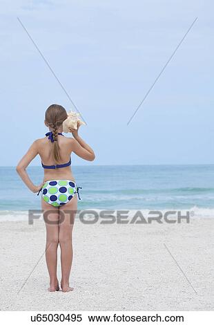 Stock Photography - USA, Florida, St. Pete Beach, Rear view of girl (8-9) listening to conch shell on beach. Fotosearch