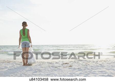 Stock Photo - USA, Florida, St. Petersburg, Rear view of girl (10-11) standing on beach with garbage bag, facing ocean. Fotosearch
