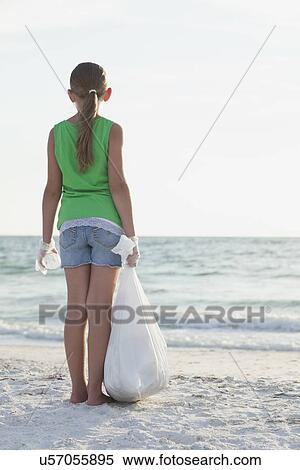 Stock Photography - USA, Florida, St. Petersburg, Rear view of girl (10-11) standing on beach with garbage bag, facing ocean. Fotosearch