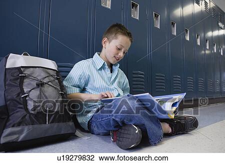 Stock Image - USA, Illinois, Metamora, Boy (8-9) sitting at lockers in school corridor and reading book. Fotosearch
