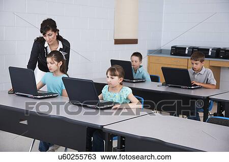 Stock Image - USA, Illinois, Metamora, Female teacher assisting children (8-9, 10-11) using laptops in computer lab. Fotosearch