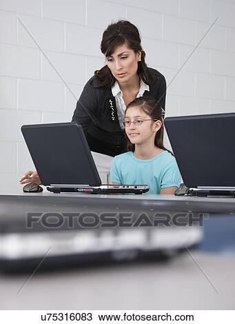 Stock Image - USA, Illinois, Metamora, Female teacher assisting girl (8-9) using laptop in computer lab. Fotosearch