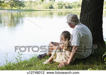 Stock Image - USA, Illinois, Metamora, grandfather and granddaughter (4-5) sitting at edge of lake. Fotosearch