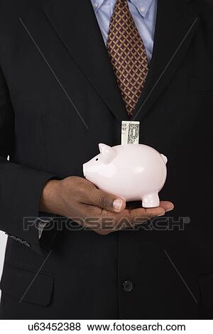 Stock Photo - USA, Illinois, Metamora, Man holding piggy bank with one dollar banknote, mid section. Fotosearch